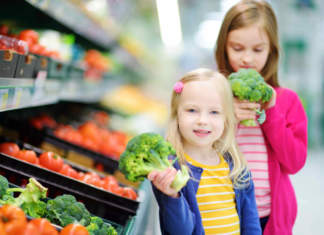 Due bambine con in mano broccoli accanto al banco degli ortaggi di un supermercato
